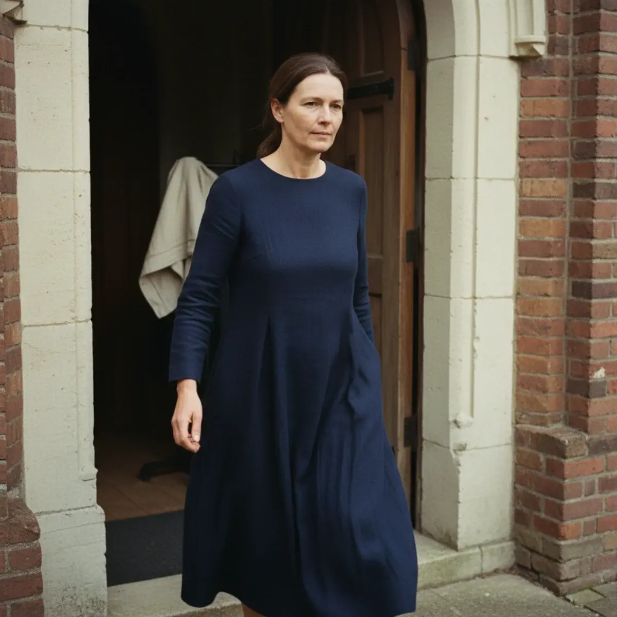 A long-sleeve navy wool midi dress with a modest neckline, paired with low-heeled tan leather pumps, opaque tights, and a small structured leather handbag