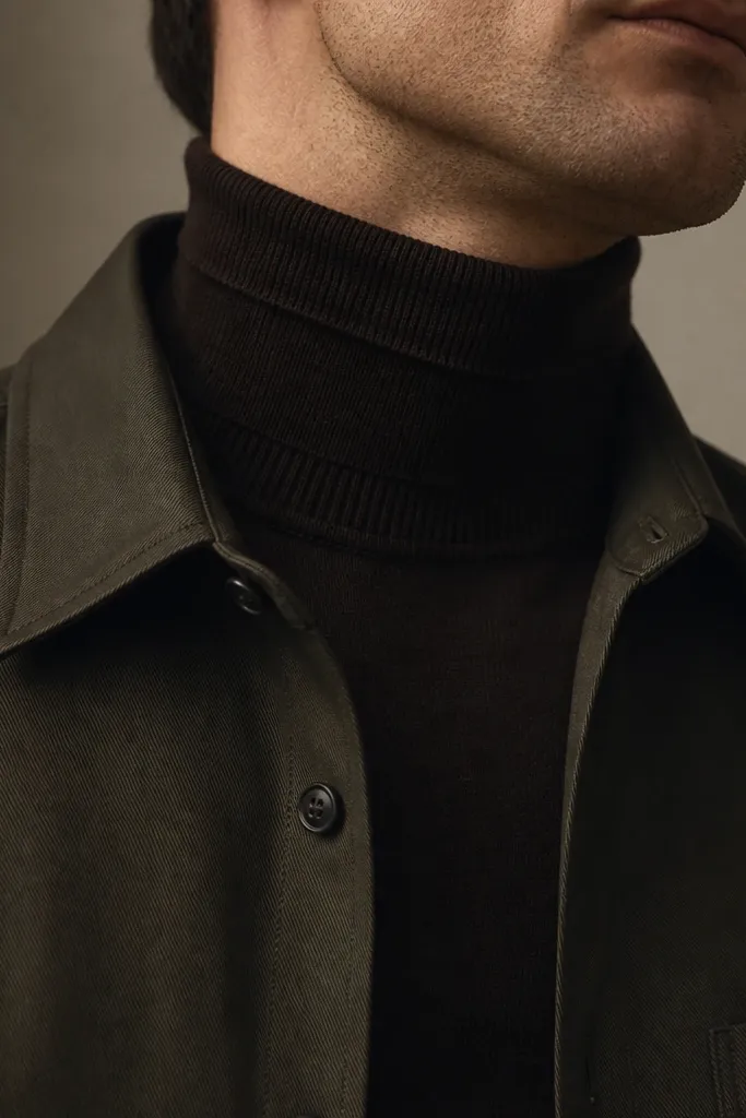 A man with a dark chocolate turtleneck under an olive overshirt, wearing black shoes and dark trousers; the photo shows the neck and collar lines.