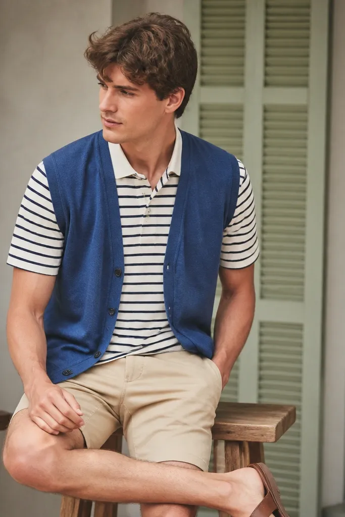 A denim-blue sleeveless cardigan layered over a striped polo shirt. The person wears beige shorts and brown sandals or loafers.