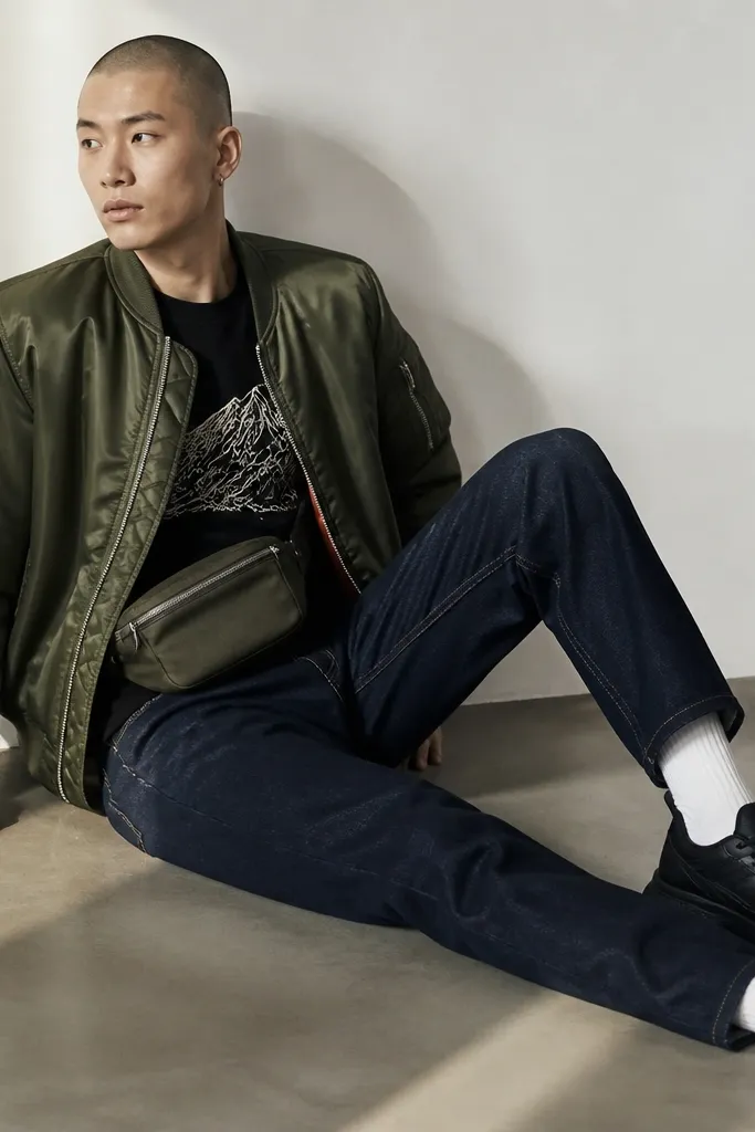A satin bomber jacket in muted olive is worn over a simple black graphic tee. The jeans are dark indigo with minimal fading. The model wears white socks, black sneakers, and a belt bag in olive or black.