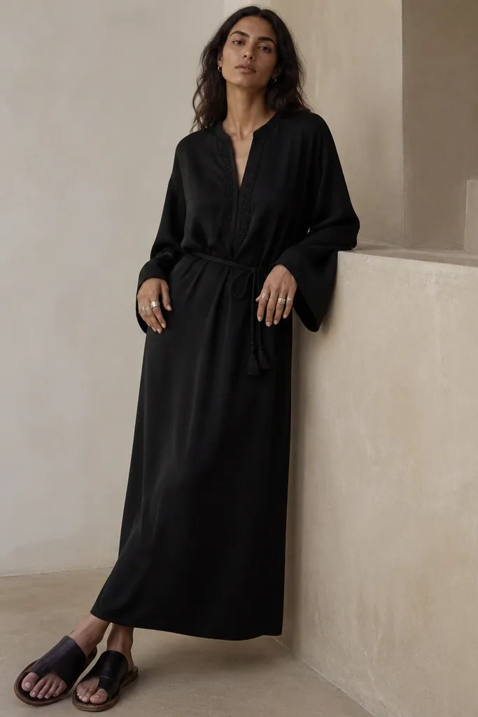 A black kaftan-style dress with a V opening, subtle embroidery on the placket, loose sleeves, and a thin black cord belt; model wears silver rings and black leather slides.