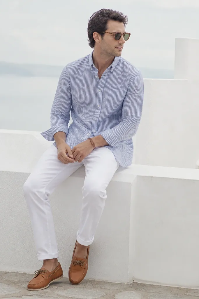 A man wearing a navy-and-white seersucker button-down shirt, paired with white chinos. He wears tan leather boat shoes and a simple braided bracelet.